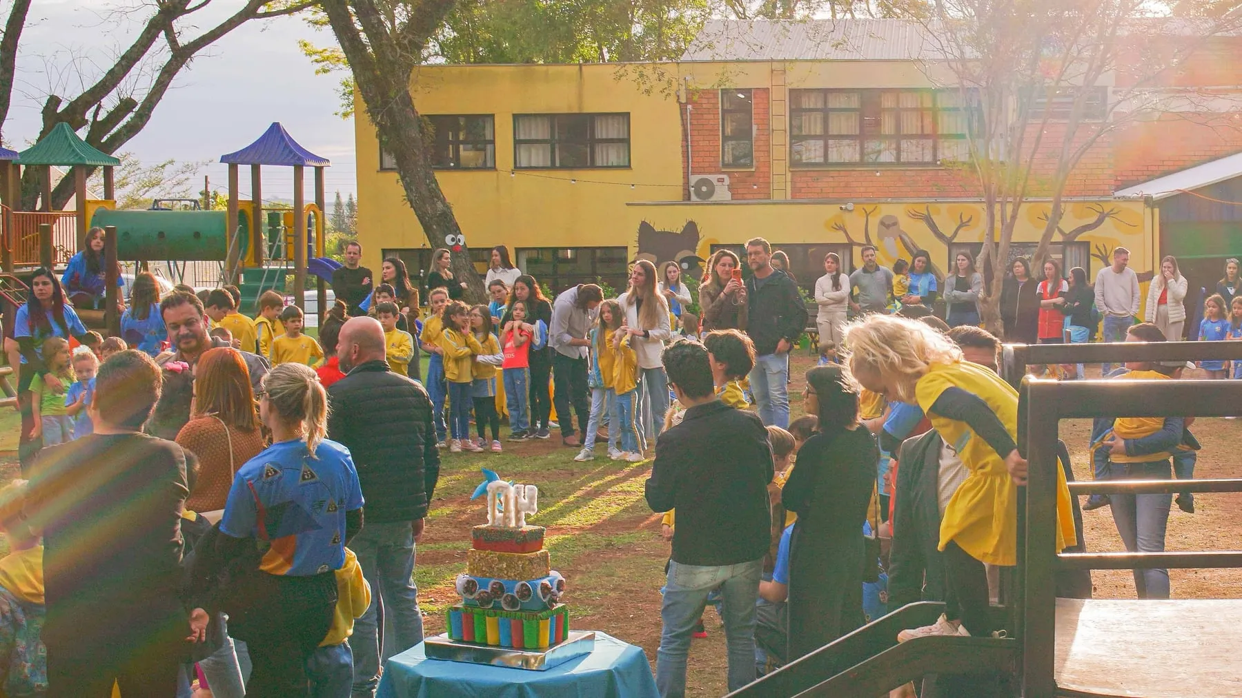 Famílias e estudantes reunidos no pátio da escola ao entardecer, em volta de uma mesa com um bolo de velas com o número 40