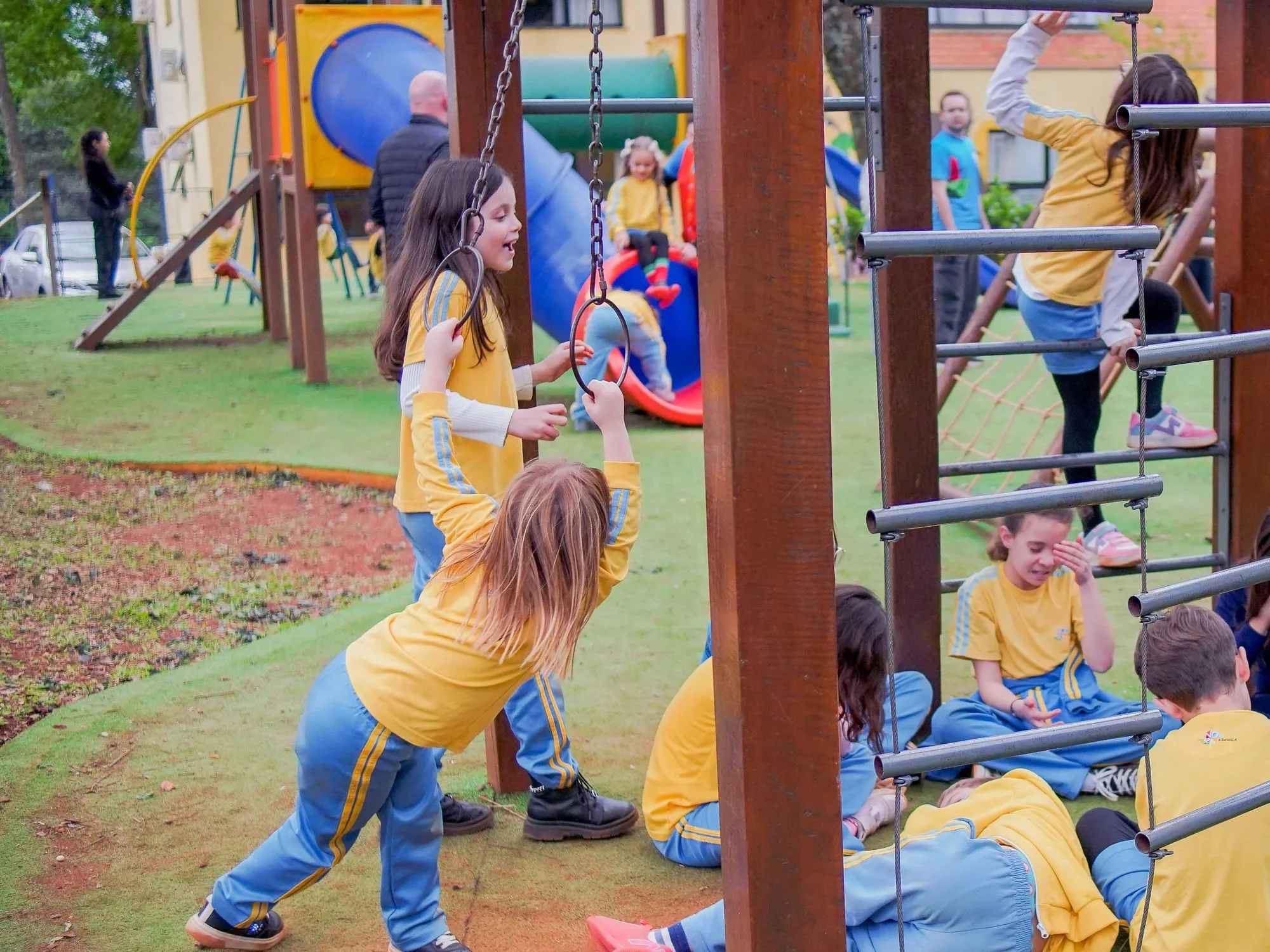 Crianças de uniforme amarelo e azul brincando nas argolas e na estrutura de escalada do parque da escola